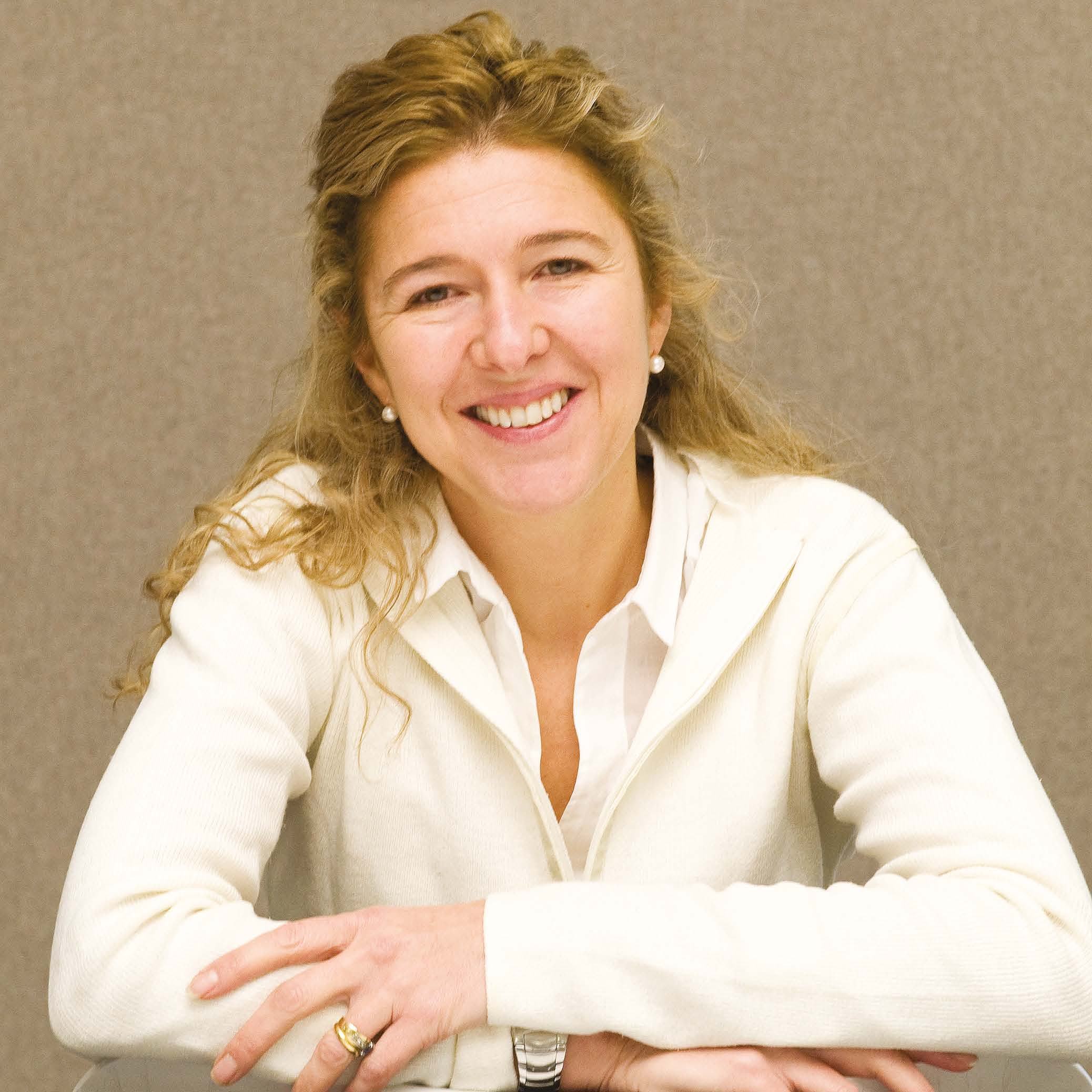 Portrait of a smiling woman with long blonde hair, wearing a white cardigan, with arms crossed on a table.