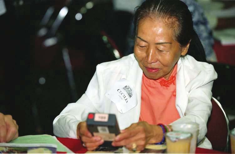 An elderly woman, wearing a "CECILIA" name tag, intently looking at a small electronic device in her hands.