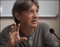 A man with short hair and a grey shirt gestures while speaking into a microphone.