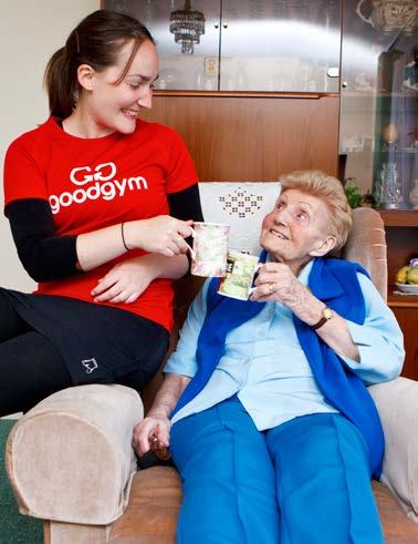 A young woman in a "goodgym" shirt shares tea and smiles with an elderly woman in an armchair, illustrating community support.