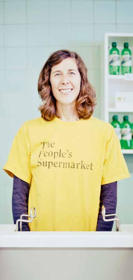 Woman in a "The People's Supermarket" t-shirt smiles behind a supermarket counter.