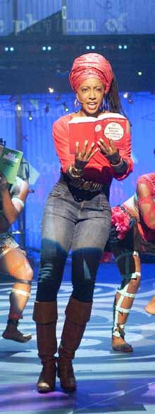 A woman in a red headwrap and shirt on stage, holding an open red book and singing, with other performers in the background.