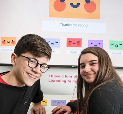 Two students smile in front of a screen displaying an emotional voting interface with various emoji-like characters representing feelings.
