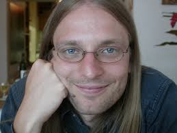 A man with long hair, glasses, and a dark shirt smiles at the camera, resting his chin on his fist.