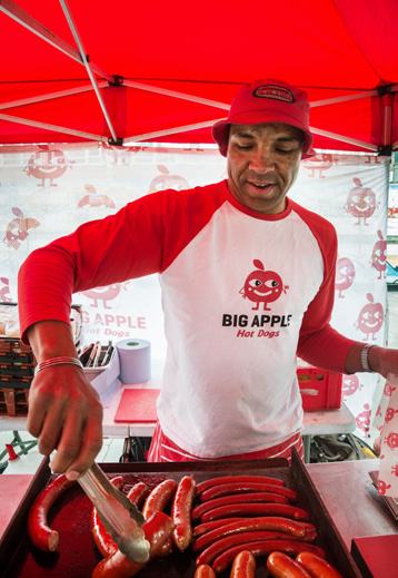 A man wearing a "Big Apple Hot Dogs" t-shirt grills sausages on a flat-top under a red tent, serving customers.