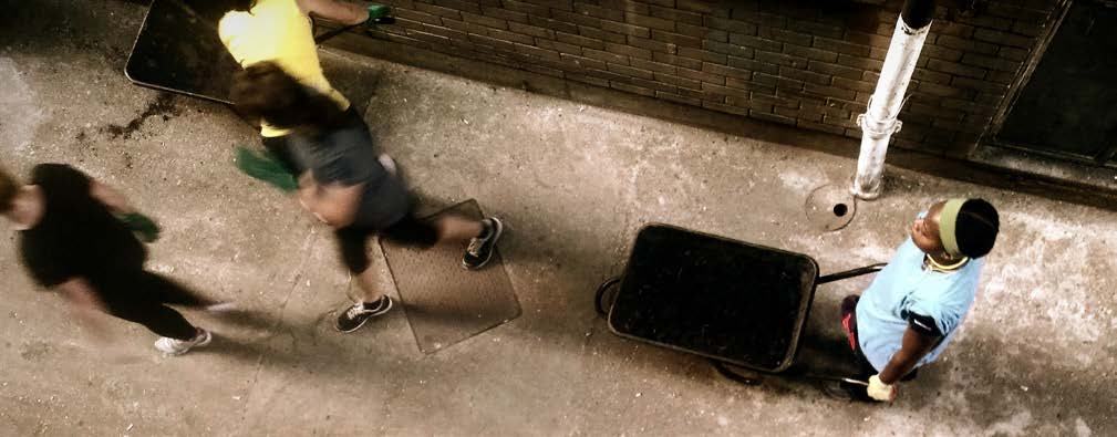 Overhead view of three people, two running quickly and one standing with a wheelbarrow, possibly engaged in an outdoor task.
