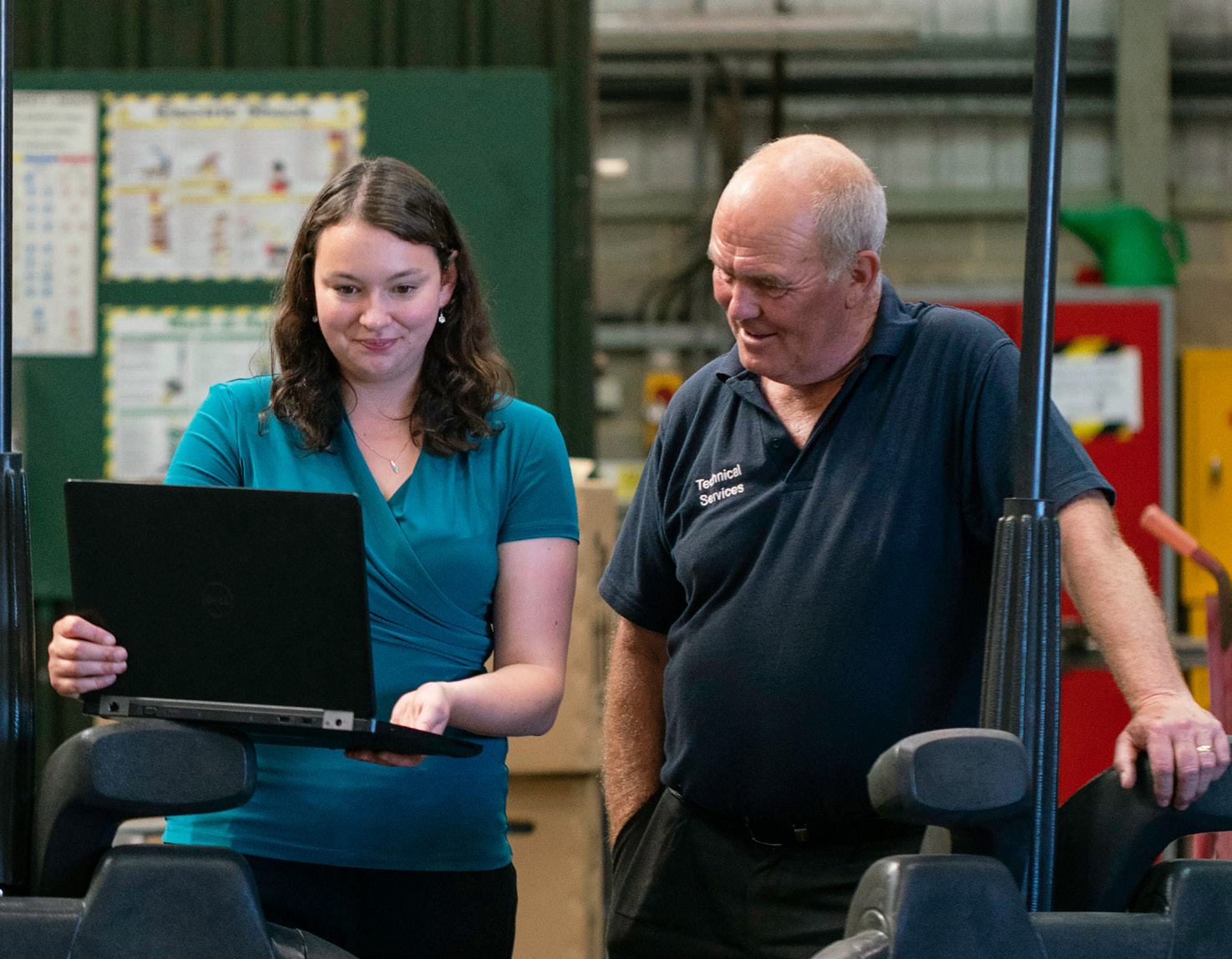 Woman showing information on a laptop to a man in an industrial setting, suggesting collaboration, training, or data review.