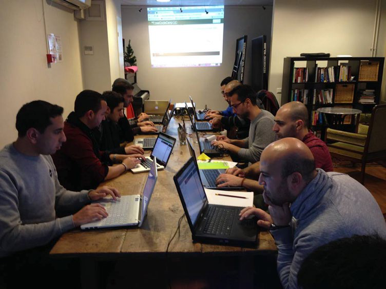 Group of people working on laptops at a long communal table, likely during a workshop or collaborative session.