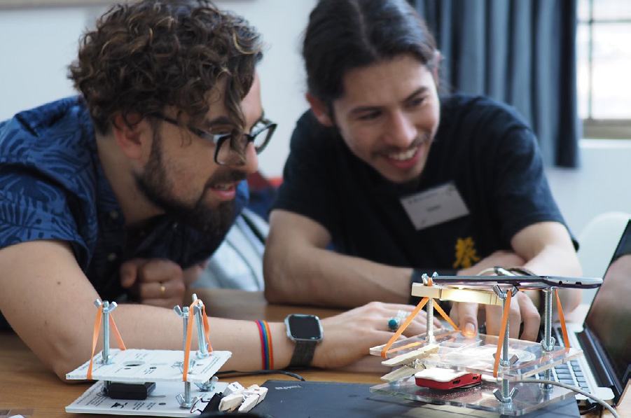 Two men examining a small, illuminated device built with clear acrylic, rubber bands, and small components, next to a laptop during a technical workshop or demonstration.