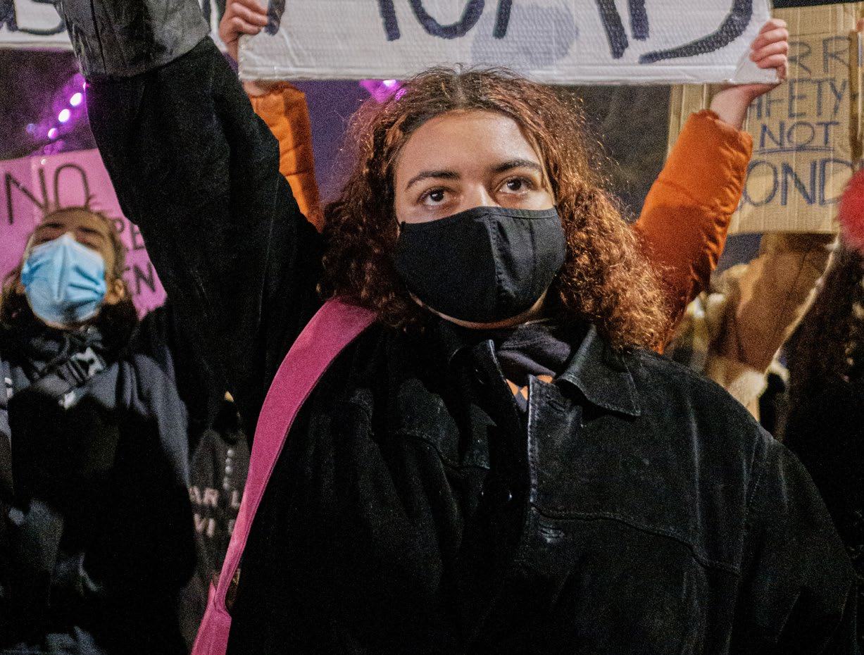 Young woman in a black face mask and jacket holding a sign at a night protest, with blurred figures in background.