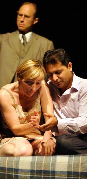 Three actors on stage; a man in a suit in the background, a man and woman in the foreground interacting on a bench.