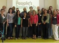 A group of adults stand together in a room, with a projector screen in the background displaying "The People's Design Lab".