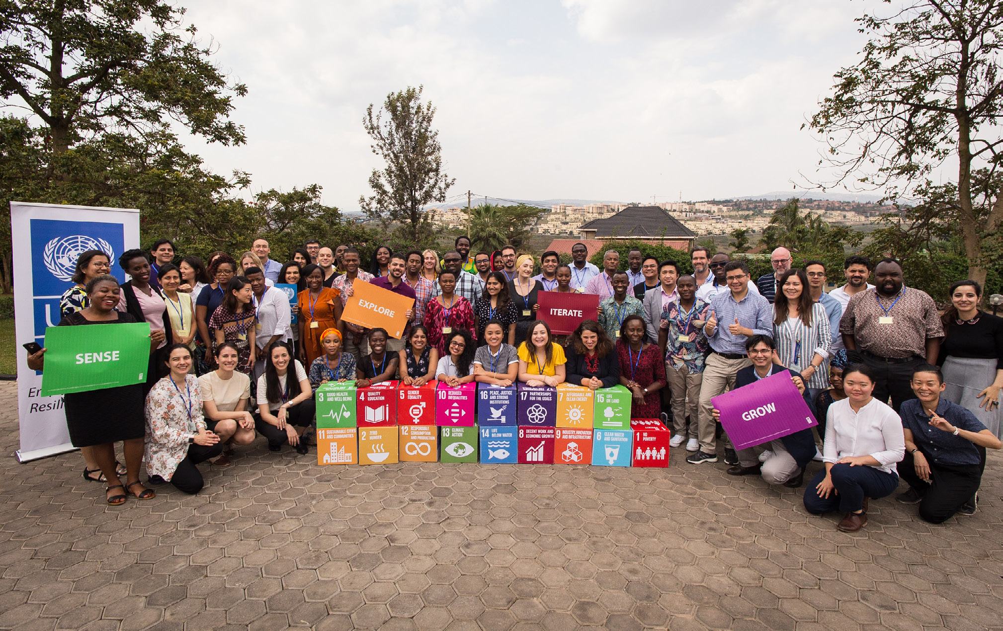 Large group of people posing with blocks displaying the 17 UN Sustainable Development Goals and signs like "SENSE", "EXPLORE", "ITERATE", "GROW".