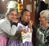 Three women in aprons, two smiling, one playfully posing with food and a book, possibly during a community cooking event.