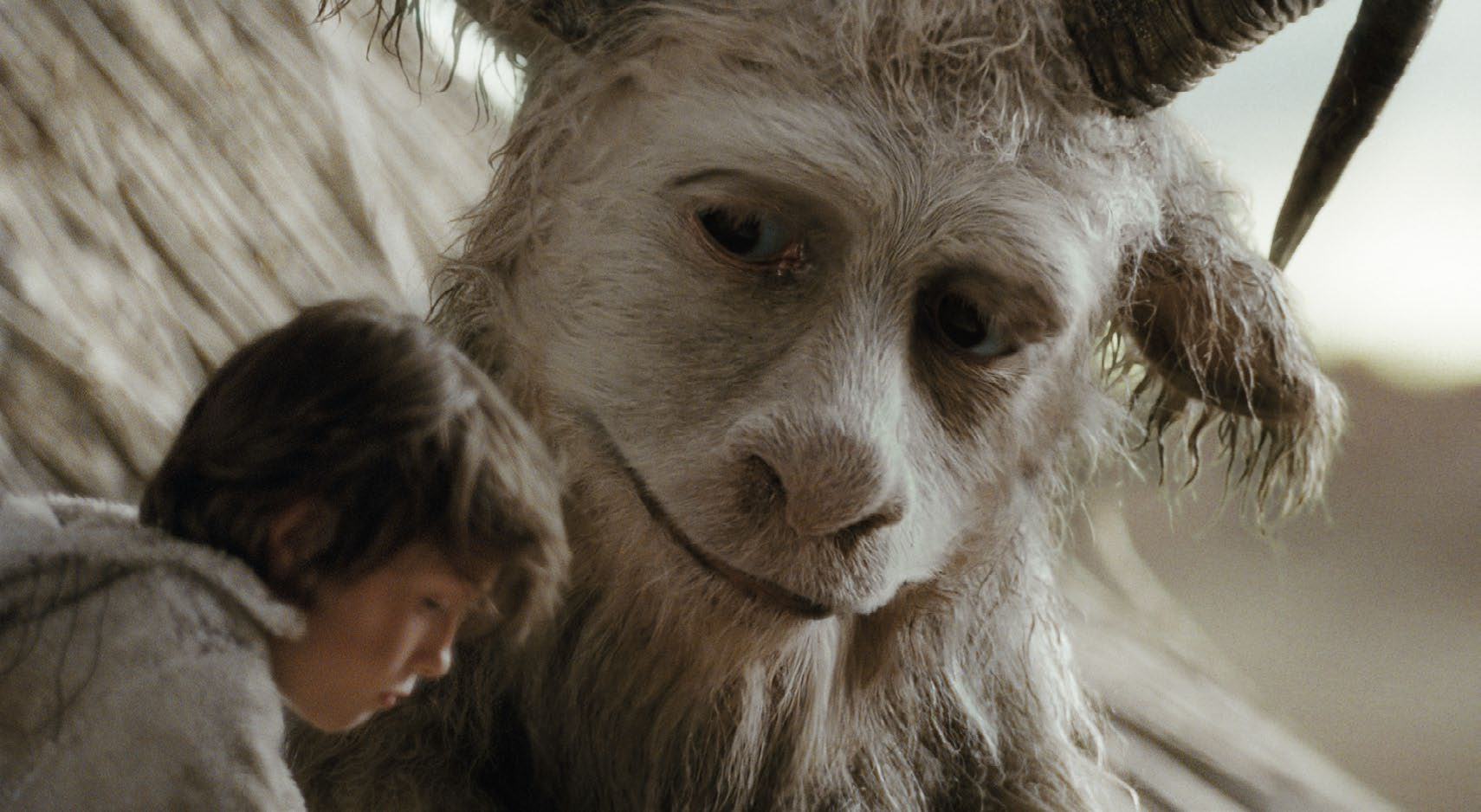 A young boy sits next to a large, furry, gentle-looking fantasy creature with small horns, conveying a moment of companionship or interaction.