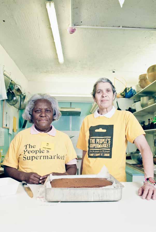 Two women in "The People's Supermarket" t-shirts stand with a cake in a kitchen.