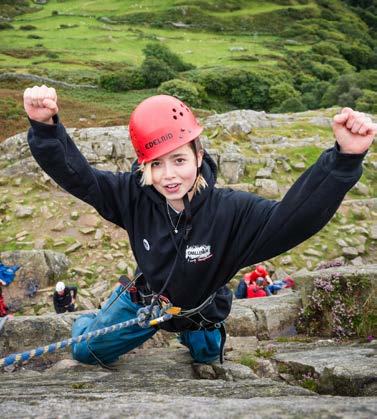 Young person in a red climbing helmet and black hoodie, harnessed to a rope, smiling and flexing on a rock face.