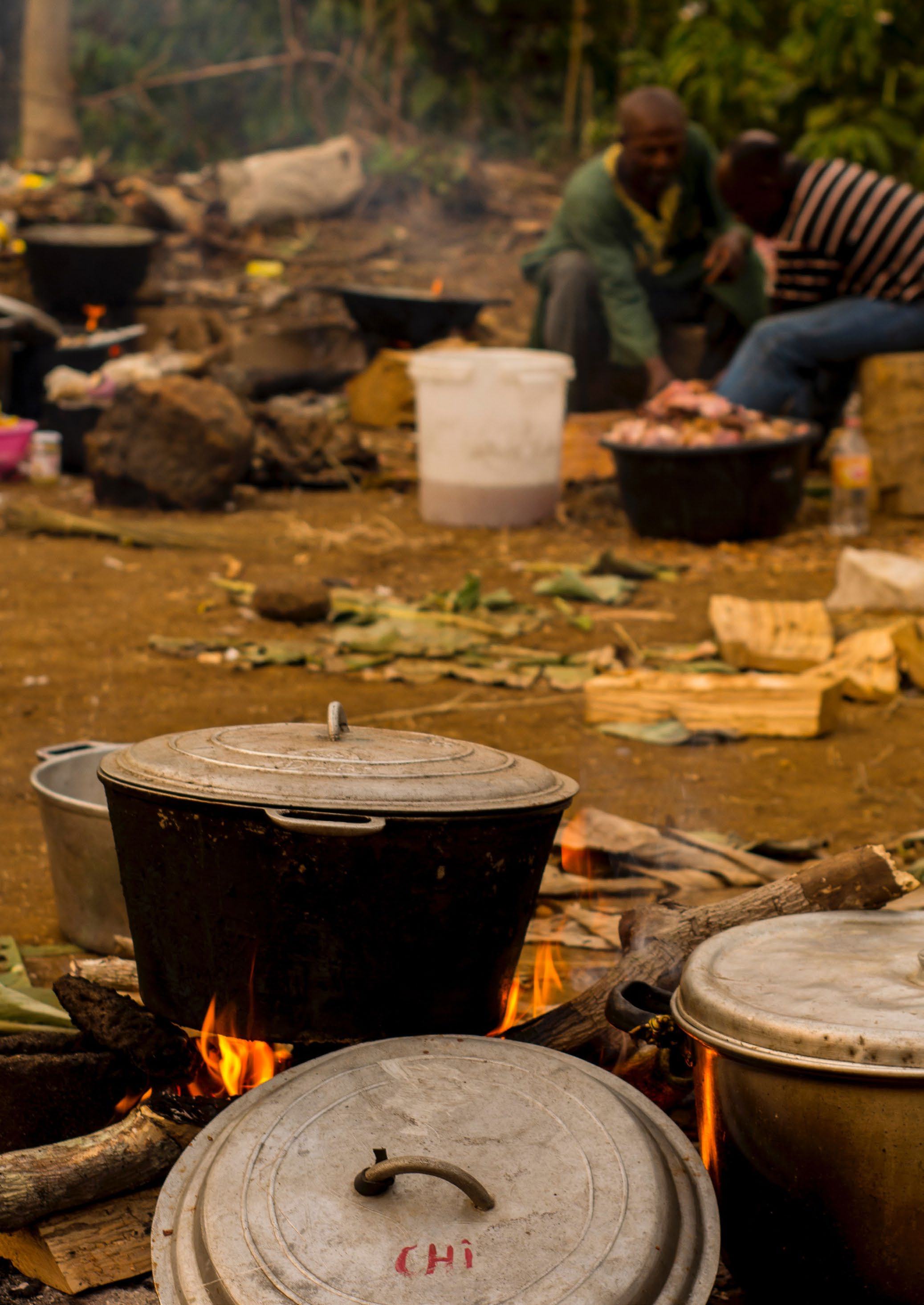 An outdoor scene showing multiple large cooking pots on open fires on the ground, with two people working in the background, depicting communal food preparation.