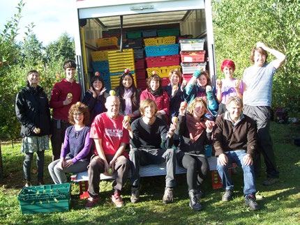 A group of people smiling outdoors with apples, standing in front of a truck loaded with crates of harvested apples.