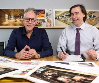 Two men, one smiling at the camera and the other writing, reviewing images and documents spread across a table.