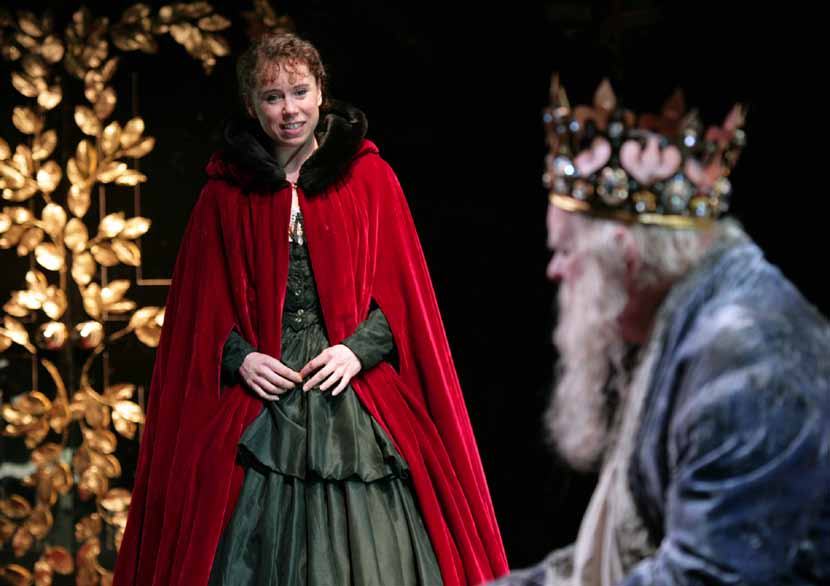 A woman in a red cape stands on stage with golden leaf decorations, while a bearded man in a crown is blurred in the foreground.