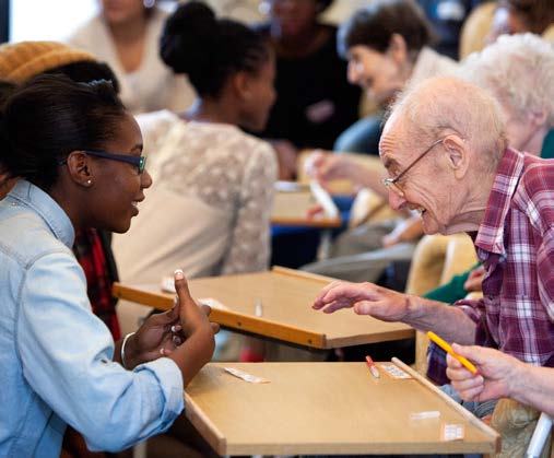 A young woman and an elderly man smile and interact across a table with small game pieces, showing intergenerational engagement.