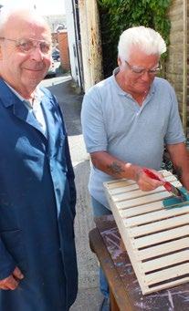 Two older men, one watching and smiling, the other painting a wooden slatted crate outdoors.