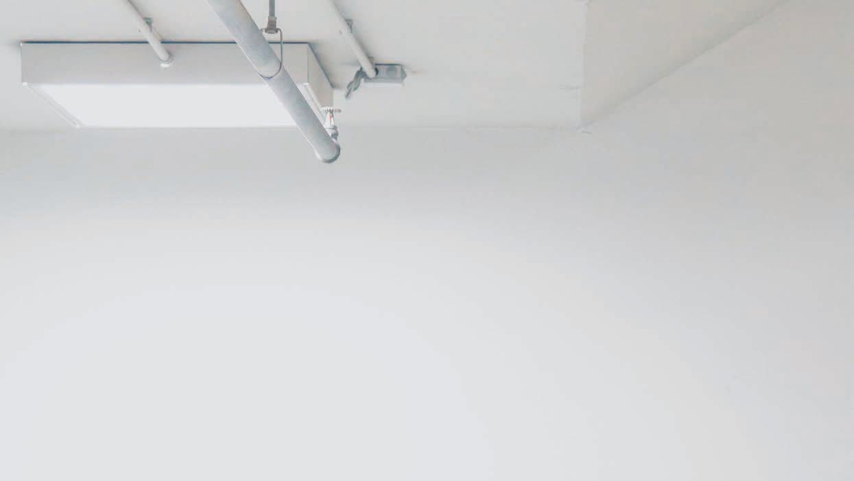 Image of a white ceiling with exposed metal pipes, fire sprinklers, and a rectangular light fixture against a plain white wall.