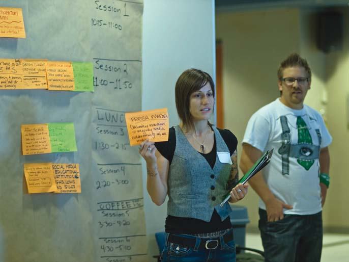 Two people, a woman holding a sticky note, standing next to a wall covered with session schedules and ideas written on sticky notes.