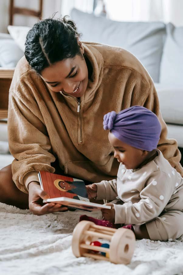 A woman and a baby in a purple turban sit on a patterned rug, looking at an open children's book together.