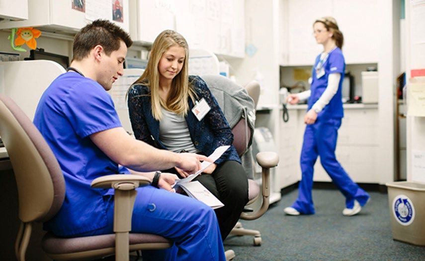 A male nurse consults with a female patient, showing her a document, in a clinical setting with another nurse walking in the background.