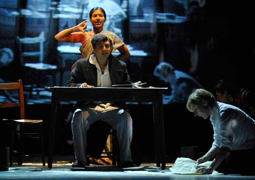 Three actors on stage; a man at a table, a woman behind him gesturing, and another person on the floor.