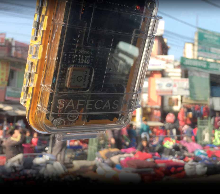 Close-up product photo of a transparent orange "SAFECAS" device case, showing an internal circuit board, with a blurred street market background.