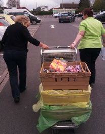 Two women from behind pushing heavily loaded shopping trolleys filled with various items across a car park.