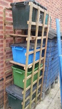 A wooden trellis shelf unit holding multiple large bins for storage or recycling next to a blue shed.