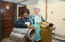 Two women in aprons smiling, standing next to large, old industrial machines, possibly in a production or processing facility.