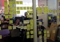 Person working behind a glass wall covered in numerous yellow sticky notes, representing brainstorming or project planning.