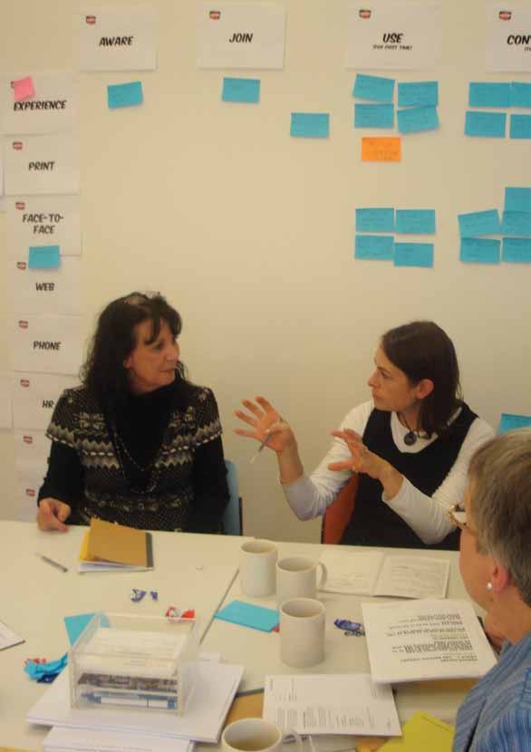 Two women in a workshop, one gesturing while talking, with a whiteboard covered in sticky notes and labels like "AWARE," "JOIN," and "USE" in the background.