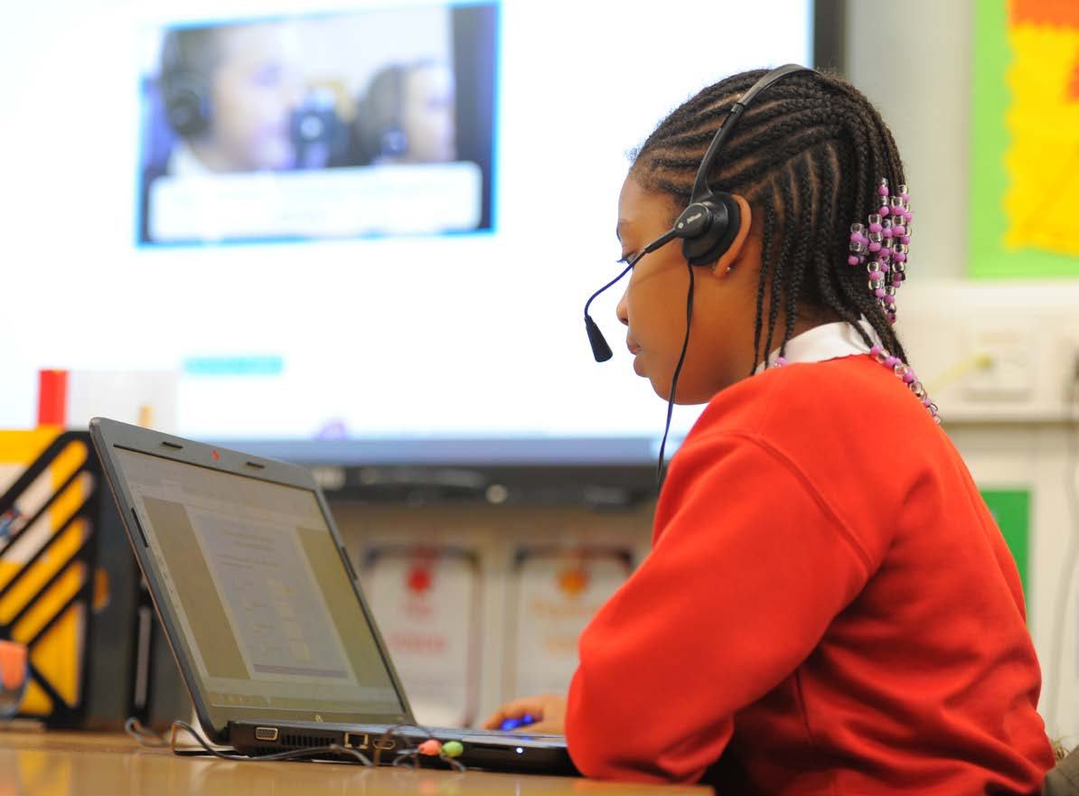 Young student with headphones and microphone using a laptop, with a video call or lesson displayed on a large screen in the background.