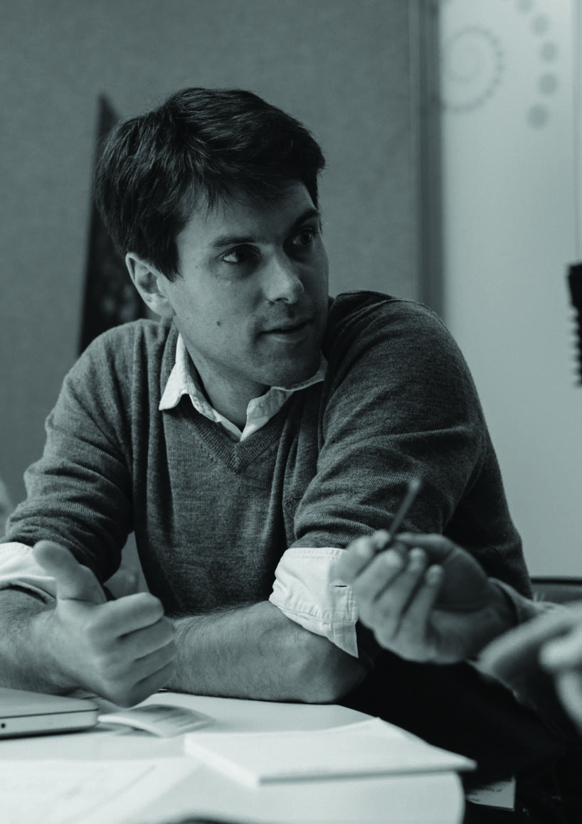 Black and white photo of a man in a sweater and collared shirt, engaged in a discussion at a table, gesturing with one hand and holding a pen.