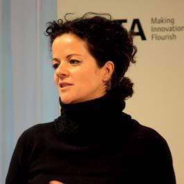 Close-up portrait of a woman with dark curly hair, wearing a black turtleneck, speaking at an event with Nesta branding.