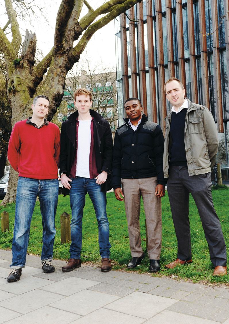 Four men standing outdoors on a paved path in front of a large tree and a modern building, possibly a team photo.