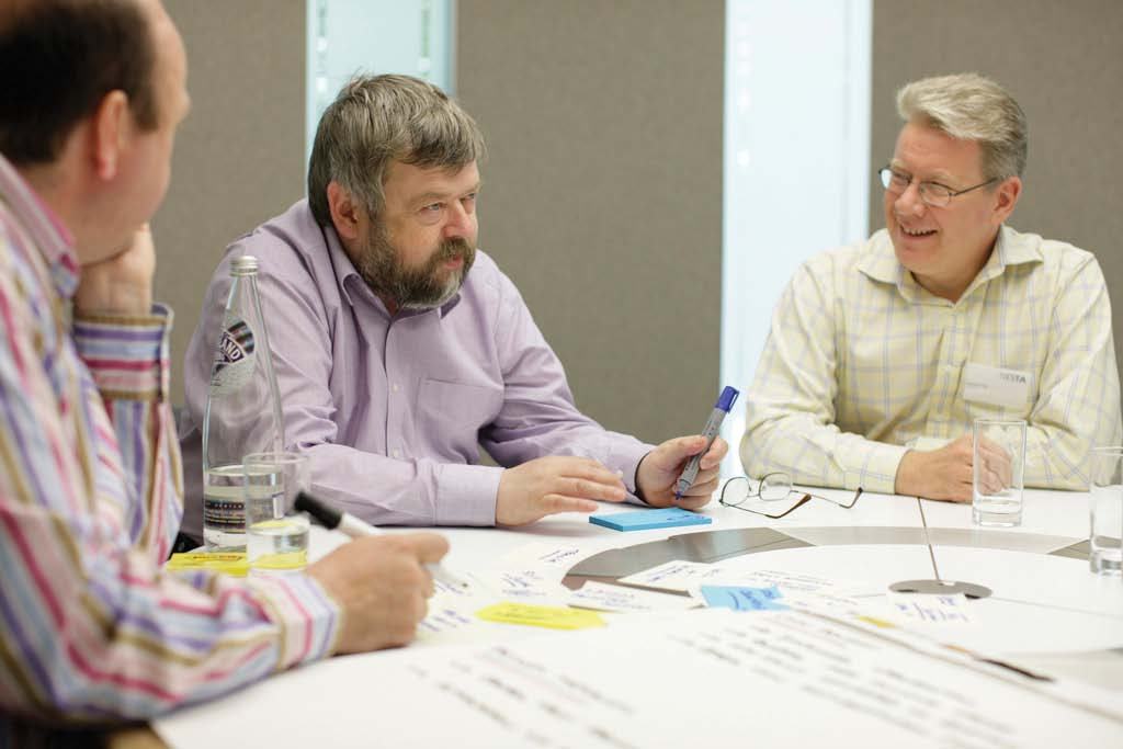 Three men at a round table discussing, with notes and markers visible on the table, indicating a collaborative work session.