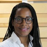 Headshot of a woman with braided hair and glasses, wearing a white shirt, smiling.