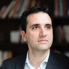 Headshot of a man with dark hair and a dark suit, looking to the right, with a bookshelf in the background.