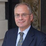 Headshot of a man with glasses, wearing a blue suit and striped tie, smiling slightly.
