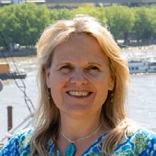 Headshot of a smiling woman with blonde hair and a patterned blue top, with a river in the background.