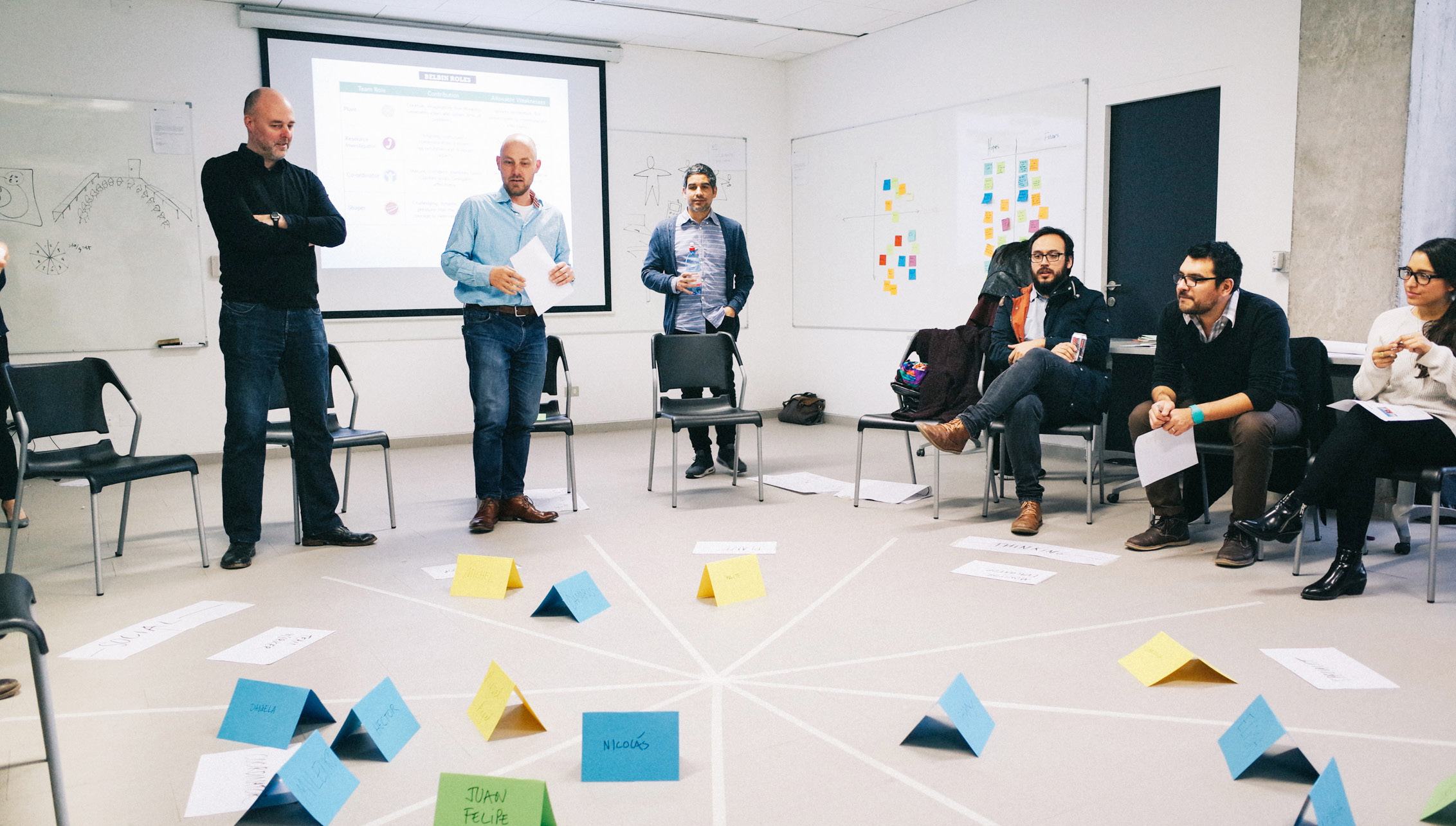 Workshop scene with people arranged in a circle, folded papers on a marked floor diagram, and a projected screen, suggesting an interactive ideation session.