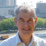 Professional headshot of an older man with graying hair, smiling, with a blurred cityscape and river in the background.