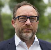 Headshot of a man with a beard and glasses, wearing a dark jacket over a white shirt, against a blurred green outdoor background.